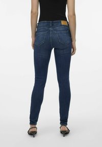 Jeans skinny azul oscuro en denim elástico, con una etiqueta de cuero marrón en la parte de atrás. Diseño de cinco bolsillos con costuras visibles y un suave desgastado.