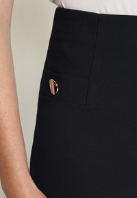 Svart högmidjad kjol med en slät textur, sidoficka med en guldaccent och dold dragkedjedetalj.