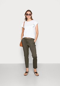 Camiseta blanca de manga corta con un gráfico de corazón, combinada con pantalones verde oliva, sandalias marrones y gafas de sol de gran tamaño. Look sencillo y casual.