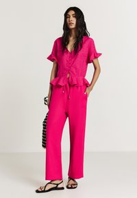 Fuchsia Peplum-Oberteil mit kurzen Rüschenärmel, mit einer Knopfleiste vorne; kombiniert mit weit geschnittenen fuchsia-farbenen Hosen und Riemchensandalen.