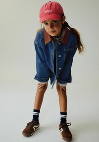 Jauna mergina dėvi raudoną „gelato“ kepurę, per dydį didelį džinsinį švarką, juodas kojines su baltais dryžiais ir rudus sportinius batelius, atsisukusi į priekį ir laikydama rankas ant kelių.