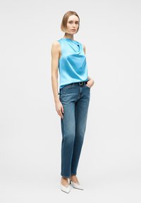 Sieviete, tērpusies bezrokas mirdzoša gaiši zila topā, taisnās zilajos džinsu biksēs ar melnu jostu un baltos augstpapēžu apavos, stāv pret vienkāršu gaišu fonu.