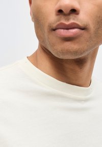 Κοντινό στην κάτω περιοχή του προσώπου και τον λαιμό ενός άνδρα που φοράει απλό λευκό μπλουζάκι με στρογγυλή λαιμόκοψη σε ανοιχτό φόντο.