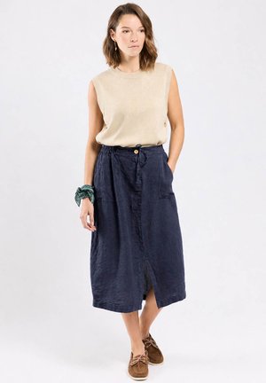 Femme portant un haut sans manches beige en tricot, une jupe midi bleu marine avec une fente devant, des chaussures marron à lacets et un chouchou vert au poignet gauche.