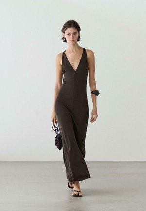 Kobieta w bezrękawniku, ciemnobrązowym kombinezonie z głębokim dekoltem w kształcie litery V, idąca boso, trzymająca małą dzianą ciemną torebkę i nosząca czarną bransoletkę na nadgarstku.