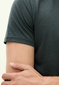 Mörkgrå t-shirt med korta ärmar, tillverkad av mjuk bomull, med en slät textur och rundad halsringning. Arm synlig, med hand vilande på armbågen.