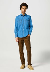 Chemise en denim bleu avec des fermetures à boutons, deux poches poitrine et des coutures de style Western ; associée à un pantalon texturé marron et des chaussures beiges.