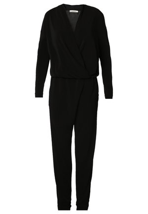 Woman wearing a black long-sleeve jumpsuit with a deep V-neck and black pointed-toe heels, accessorized with a gold pendant necklace.