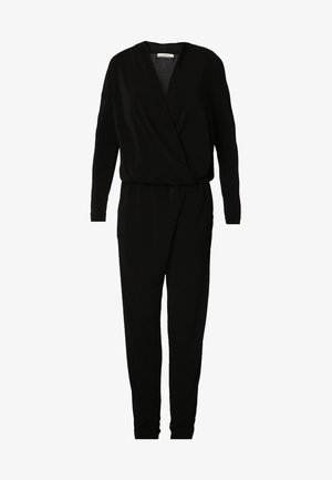 Femme portant une combinaison noire à manches longues avec un décolleté en V profond et des escarpins noirs à bout pointu, accessoirisée d'un collier pendentif en or.