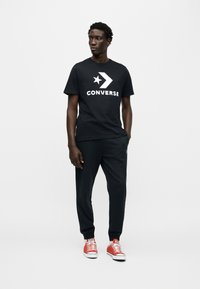 Μαύρο βαμβακερό t-shirt με λευκό λογότυπο "Converse" και αστέρι, συνδυασμένο με μαύρη φόρμα και κόκκινα αθλητικά παπούτσια με λευκές λεπτομέρειες.