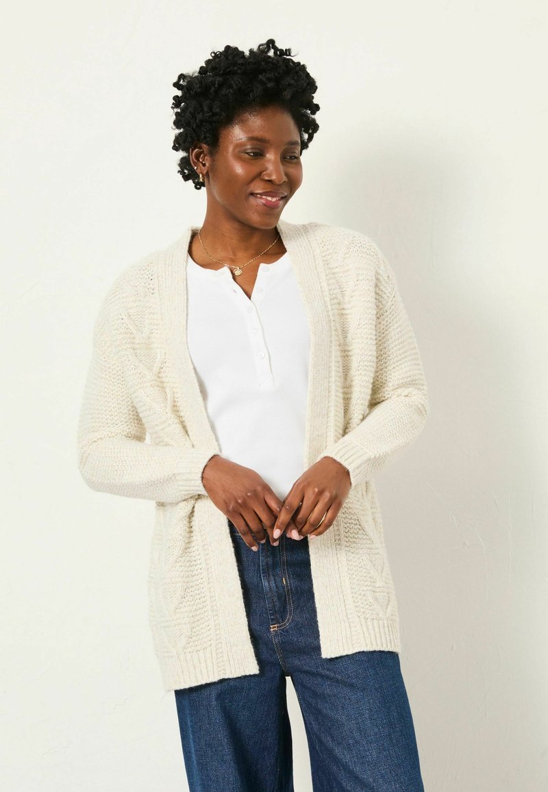 Strickjacke in Creme mit langen Ärmeln, offener Vorderseite und strukturierter Musterung, getragen über einem weißen Hemd und kombiniert mit dunkelblauen Jeans.