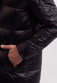 Černá péřová bunda s prošívan�ými částmi, nášivkou s logem na rukávu a bočními kapsami. Materiál vypadá hladký a lesklý.