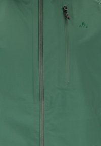 Vihreä takki, jossa pystysuuntainen etuvetoketju, rintataskussa vetoketju ja vetoketjynvetimen reuna sekä pieni kolmionmuotoinen logo rinnassa.