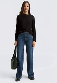Black long-sleeve sweater with round neckline, paired with blue wide-leg jeans. Smooth texture; model holding a dark green handbag.