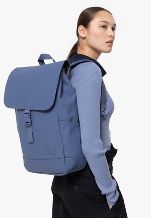 Mochila azul con cierre de solapa, hecha de material suave. Cuenta con correas ajustables, hebilla frontal y bolsillos laterales.