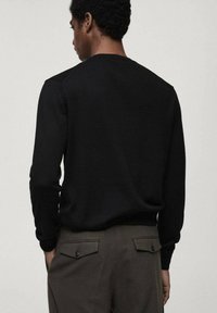 Schwarzer Pullover mit langen Ärmeln, geripptem Ausschnitt und Saum. Weicher Stoff, taillierte Form, getragen mit dunkelgrünen Hosen mit Gesäßtaschen.