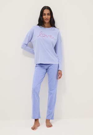 Woman standing barefoot wearing light purple long-sleeve top with "love" text and matching pants with small red heart pattern.