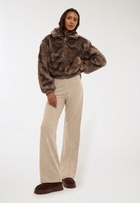 Giacca corta in finta pelliccia marrone con cappuccio, pantaloni beige a coste a gamba larga e scarpe marroni slip-on. Texture morbida e vestibilità rilassata.