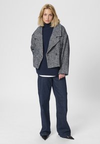 Blondinka v črno-belem jaknu v ribji kost, mornarsko modri pulover s turtleneck ovratnikom, širokih hlačah in črnih špičastih čevljih stoji ob belem ozadju.