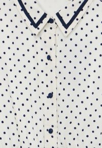 Blouse en tissu blanc avec des pois bleu marine, un col contrastant bleu marine et des boutons sur le devant. Texture lisse.