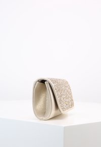 Lille guld clutch-pung med en glitrende klap, der står oprejst på en hvid overflade mod en ensfarvet baggrund.