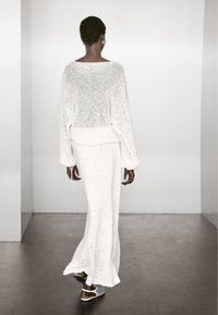 Kobieta z krótkimi włosami, ubrana w biały dziergany sweter i długą spódnicę, spaceruje po ciemnej podłodze w minimalistycznym, jasnym pomieszczeniu.