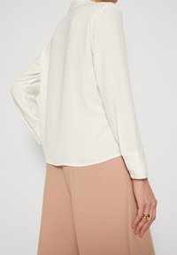 Bílá blůza s dlouhými rukávy, kulatým límcem, měkkou tkaninou, viditelnými manžetami na knoflíky a uvolněným střihem. Spárováno s taupe širokými kalhotami.