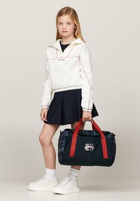 Felpa bianca con cappuccio e cuciture rosse, gonna plissée blu, sneakers bianche, mentre tiene una borsa a tracolla a scacchi blu con cinghie rosse e logo.