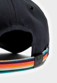 Baseball kapa v modrem bombažnem twillu s pisanimi prugastimi trakom in črnimi kovinskimi elementi. Ima ukrivljen rob in ventilacijske luknje.