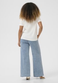Witte t-shirt met korte mouwen gecombineerd met lichtblauwe wijdlopende jeans. Model staat met de rug naar de camera, waarbij een ontspannen pasvorm en een soepele textuur worden getoond.