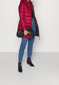 Persona vestida con una chaqueta de plumas roja, jeans azules, botas negras hasta el tobillo, sosteniendo un gran bolso de mano negro texturizado contra un fondo sencillo.