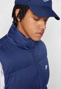 Gilet imbottito blu navy con colletto alto, chiusura con zip e un piccolo logo Nike bianco sul petto. Texture liscia, materiale leggero.
