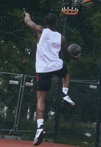 Joueur de basketball en maillot sans manches blanc et en shorts noirs, sautant pour dunker un ballon. Porte des chaussures noires avec des accents blancs et rouges.
