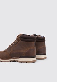 Botas de tobillo de cuero marrón con un acabado texturizado, que cuentan con un acolchado en el collar, forro de cuadros y una suela blanca contrastante.