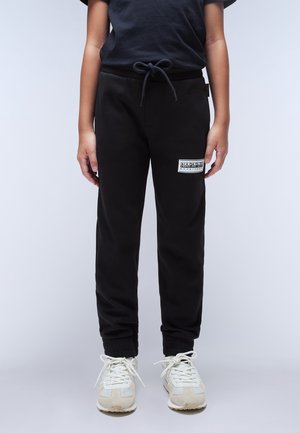 Kind trägt schwarze Jogginghose mit Kordelzug in der Taille und weiße Turnschuhe, steht vor einfachem hellem Hintergrund.