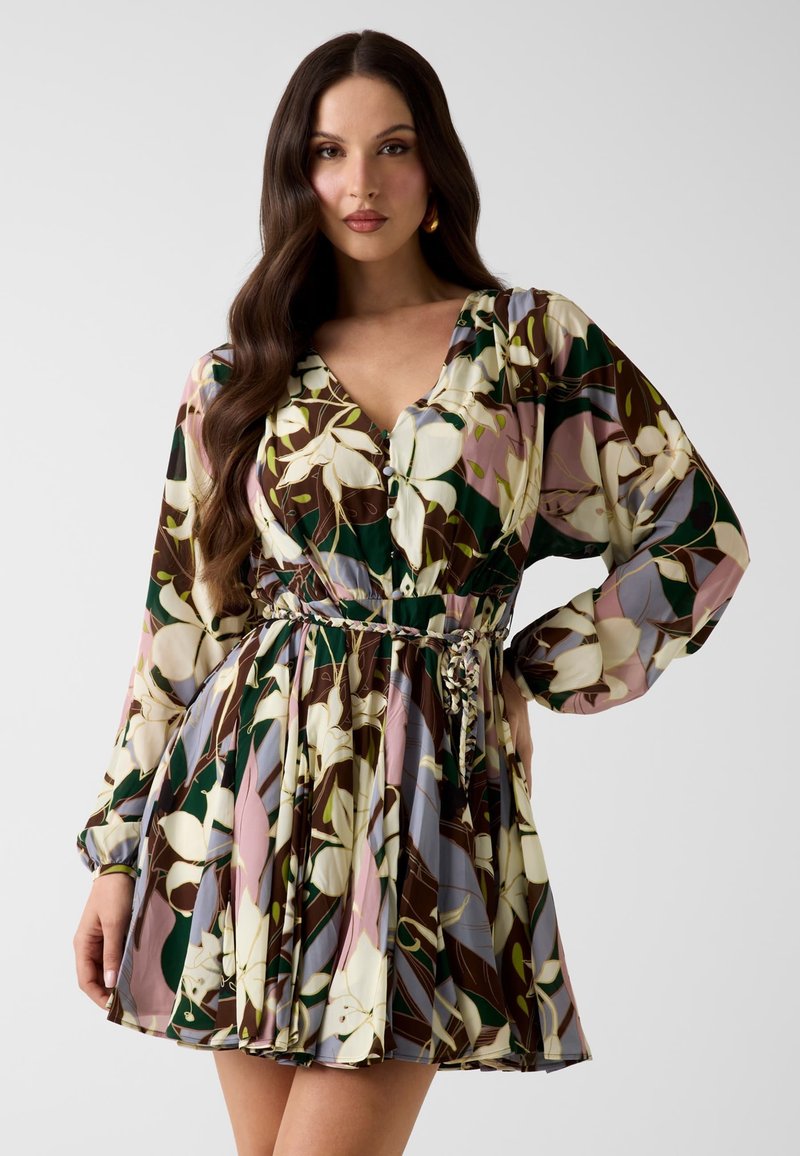Femme portant une robe à manches longues à motif floral, un col en V et une ceinture tressée, posant avec une main sur la hanche sur un fond blanc.