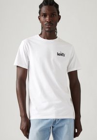 Weißes Baumwoll-T-Shirt mit kurzen Ärmeln, das ein schwarzes Levi's-Logo auf der Brust präsentiert. Der Schnitt ist lässig und wird mit hellblauen Jeans kombiniert.