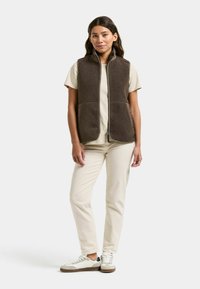 Vrouw staand, gekleed in een bruin fleecevest over een beige t-shirt, met beige broek en witte sneakers, tegen een effen achtergrond.