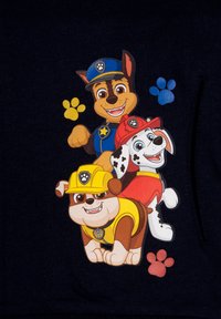 Tre cuccioli di cartone, vestiti con abiti blu, rossi e gialli, si trovano su uno sfondo blu scuro. Dettagli notevoli includono impronte di zampe in giallo e blu.