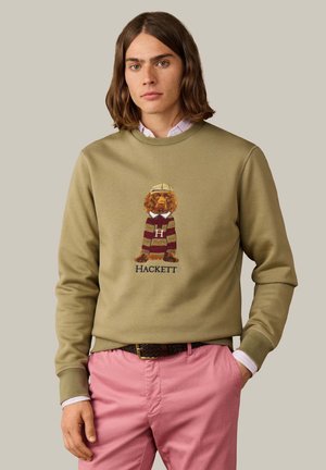 Young man with shoulder-length hair wearing an olive sweatshirt featuring a dog in a striped shirt and pink trousers with one hand in pocket.