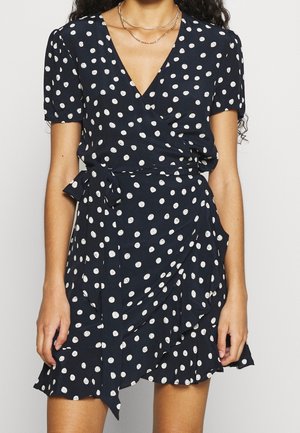 Femme portant une robe portefeuille bleu marine à pois blancs et manches courtes, accessoirisée avec des colliers en or superposés.