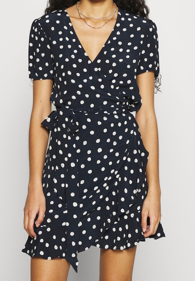 Femme portant une robe portefeuille bleu marine à pois blancs et manches courtes, accessoirisée avec des colliers en or superposés.