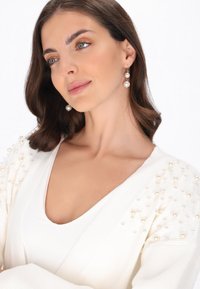 Biała dziergana kardiganka z ozdobami perłowymi na jednym ramieniu, zestawiona z białą bluzką. Gładka faktura, dekolt w kształcie litery V oraz naturalny makijaż.