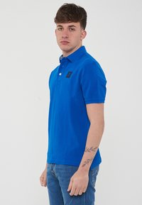 Polo shirt in cotone blu con maniche corte, colletto e una toppa nera con logo sul lato sinistro del petto. Indossato con jeans blu, evidenziando un look casual.