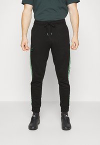 Mand iført sorte joggers med grønne striber på siderne, sorte Adidas-sko, står imod en ensfarvet lys baggrund.