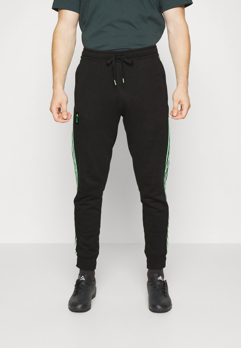 Mand iført sorte joggers med grønne striber på siderne, sorte Adidas-sko, står imod en ensfarvet lys baggrund.