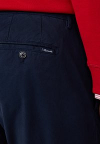 Los pantalones de algodón azul marino presentan un diseño elegante con detalles cosidos, un bolsillo trasero y un botón metálico redondo. Etiqueta de la marca visible.