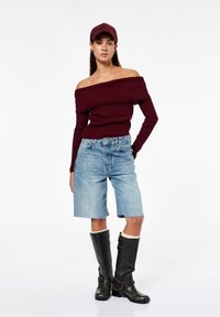 Burgunderfarbener Ripp-Strickpullover mit schulterfreier Ausführung, hellblaue Jeansshorts und knielange schwarze Stiefel mit Schnallen. Das Modell trägt eine passende burgunderfarbene Mütze.
