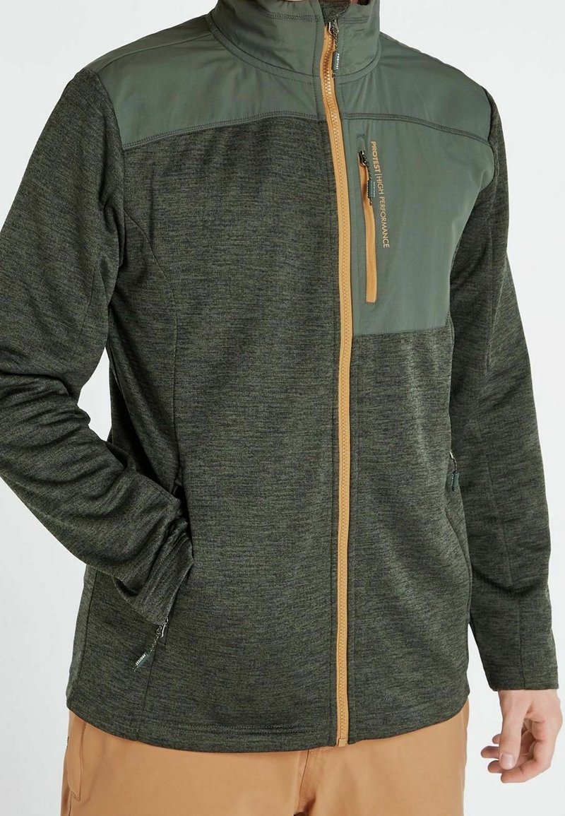 Homme portant une veste zippée vert foncé avec un panneau contrasté sur le haut de la poitrine et une poche verticale zippée, associée à un pantalon marron clair.