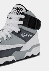 Baskets montantes en blanc, gris et noir avec des lacets blancs, une sangle noire à la cheville et la signature « Patrick Ewing » sur le côté.
